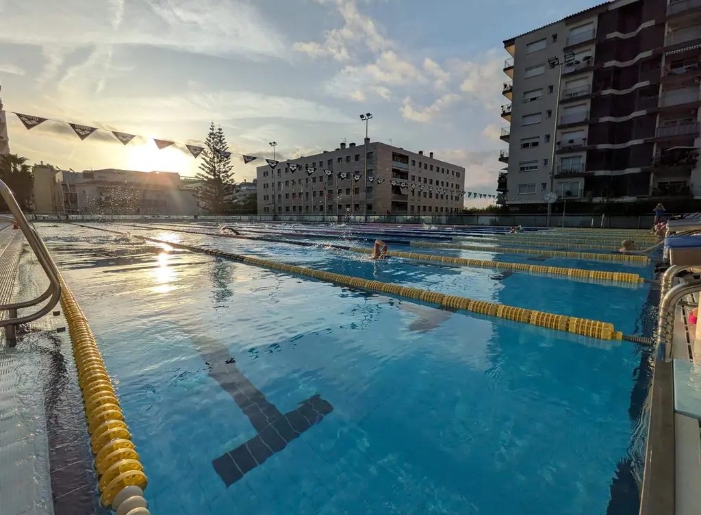 A fifty metre outdoor pool at sunrise, lane ropes running to the far end, one swimmer mid stroke with an arm extended in front.