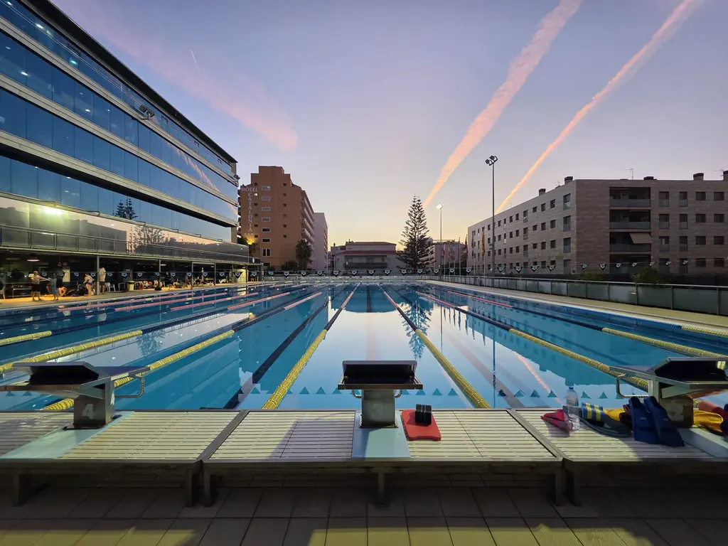 An empty fifty metre pool at first light, starting blocks in the foreground, flat water, hotel buildings behind.