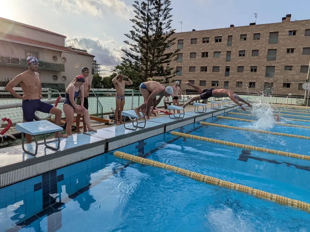 A swimmer entering the water with a splash while others stand on the poolside watching the line.