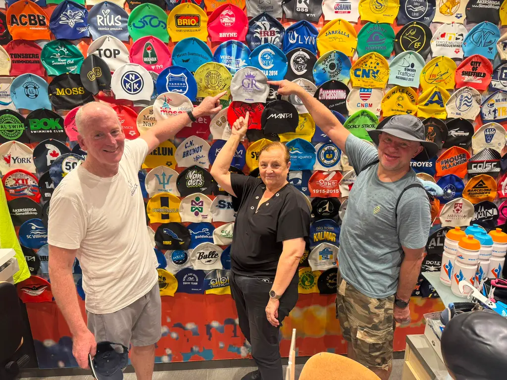 Two swimmers and a shop worker in front of a wall covered in hundreds of swimming caps from clubs around the world, one cap being lifted into place near the top.
