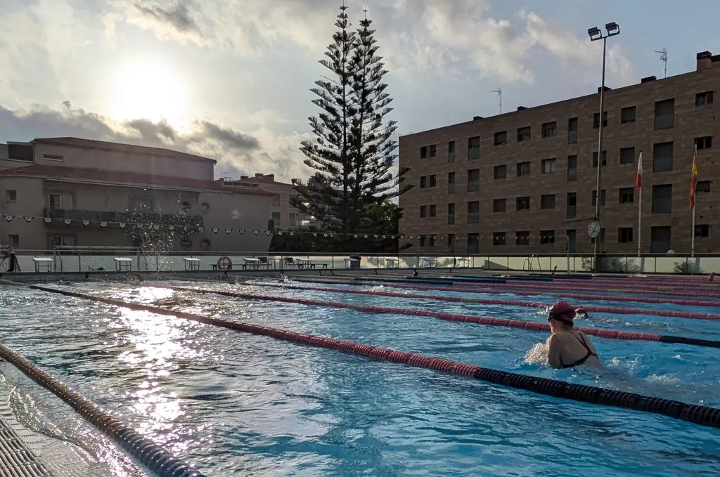 A single swimmer in a red cap in one lane, early sun flaring off the water surface.