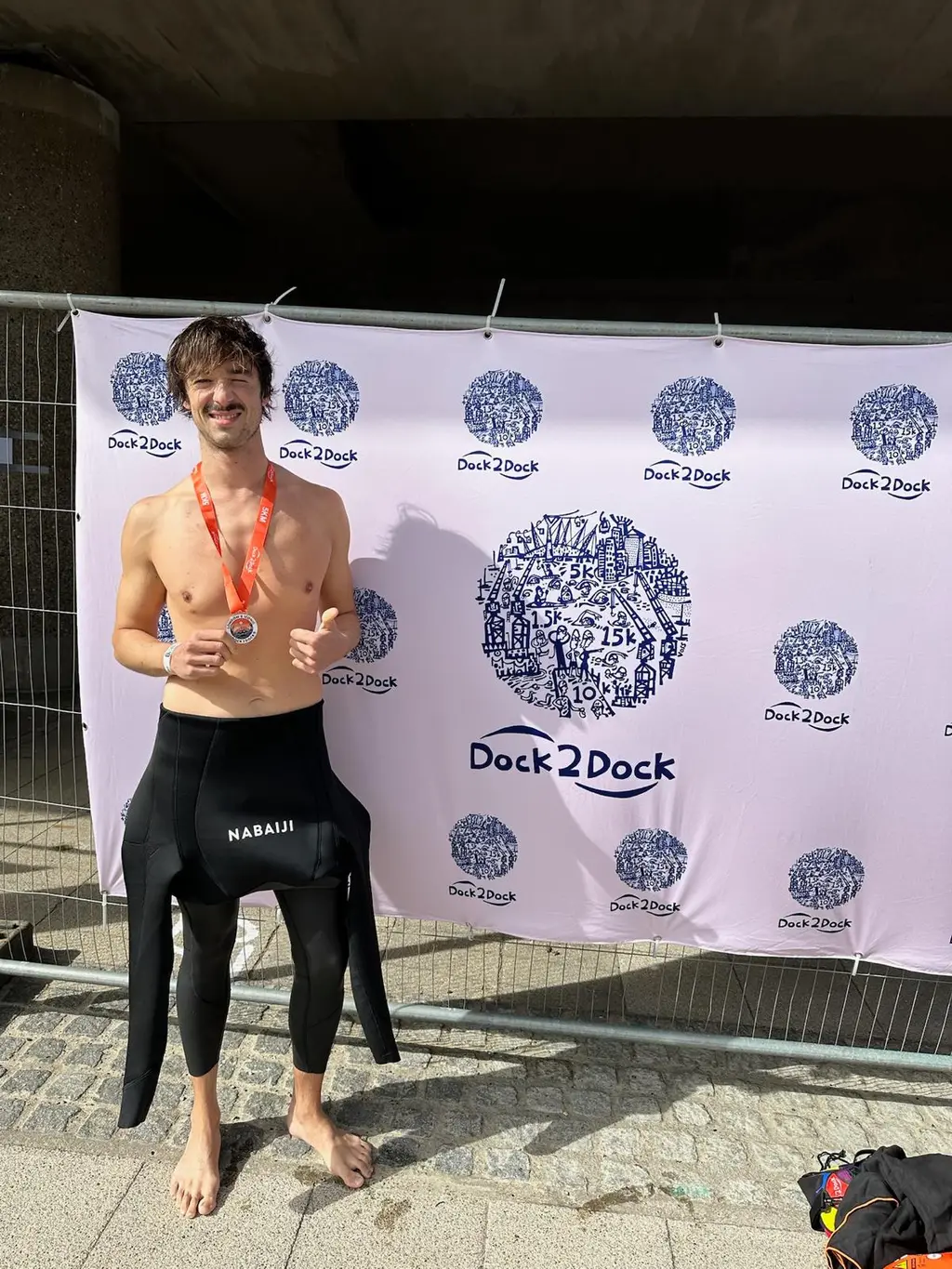 A swimmer with a medal round their neck, wetsuit peeled to the waist, one thumb up and the medal held in the other hand, in front of a white Dock2Dock banner under a concrete flyover.