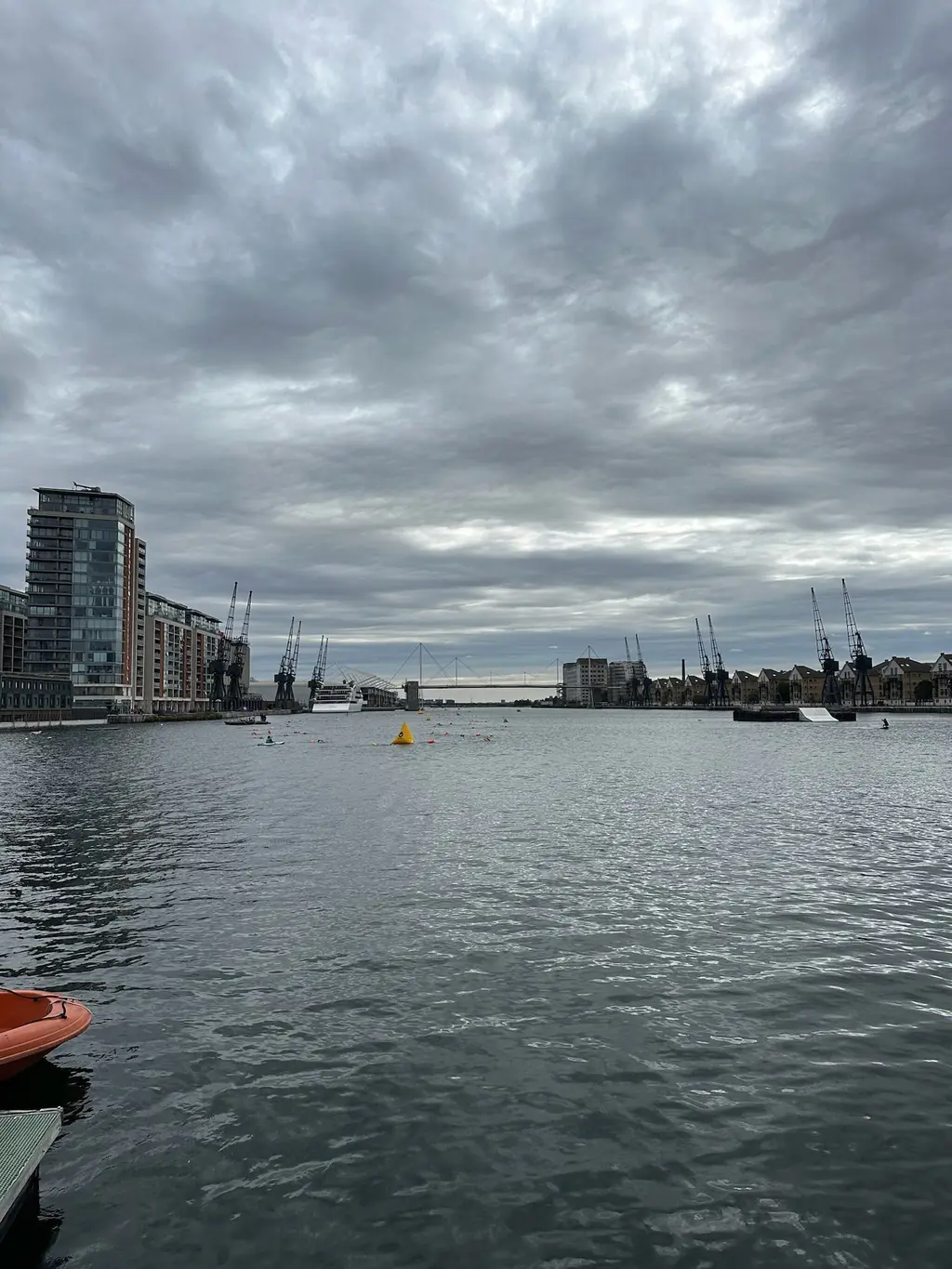 A long stretch of grey dock water under a heavy sky, apartment blocks on the left, cranes far off on the right.