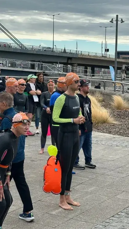 Swimmers in wetsuits and orange caps standing in a loose group on the dockside, goggles up, tow floats clipped on.