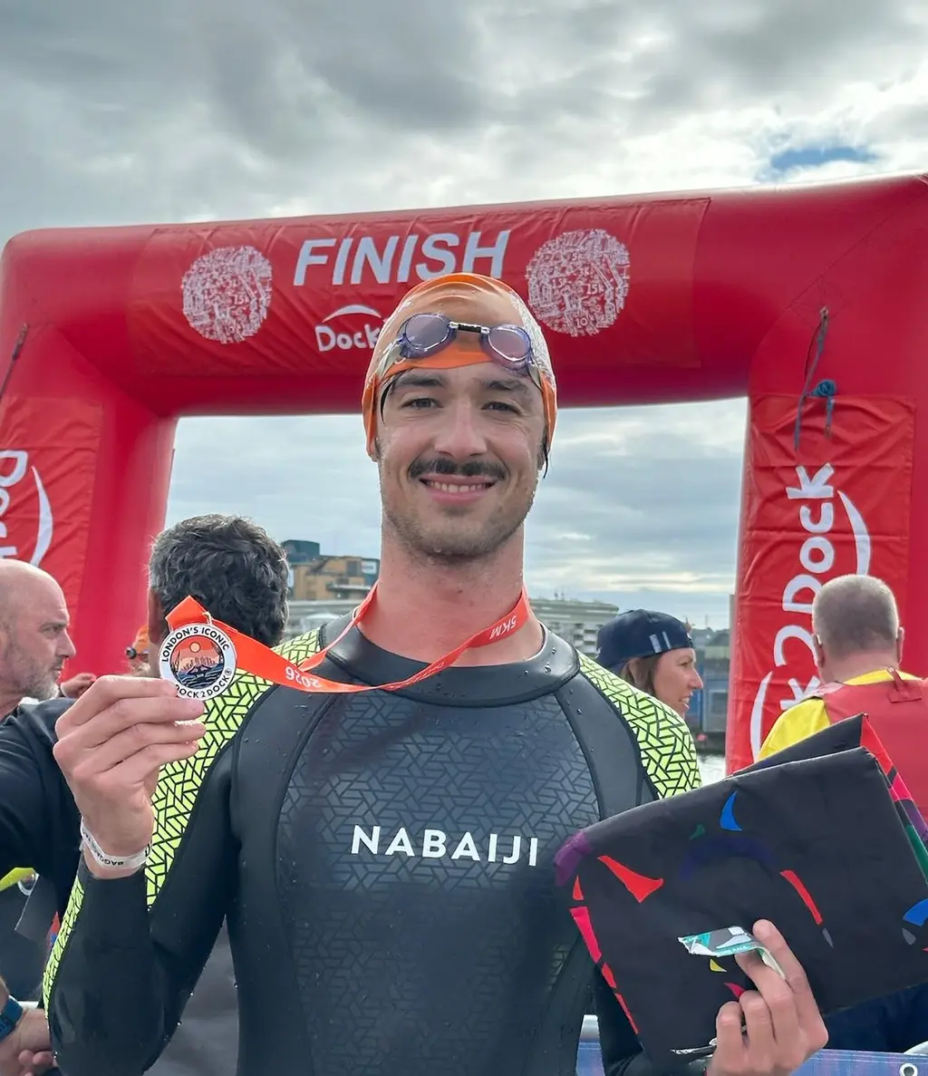 A swimmer in a wetsuit and orange cap under a red inflatable finish arch, holding up a medal, other finishers behind.