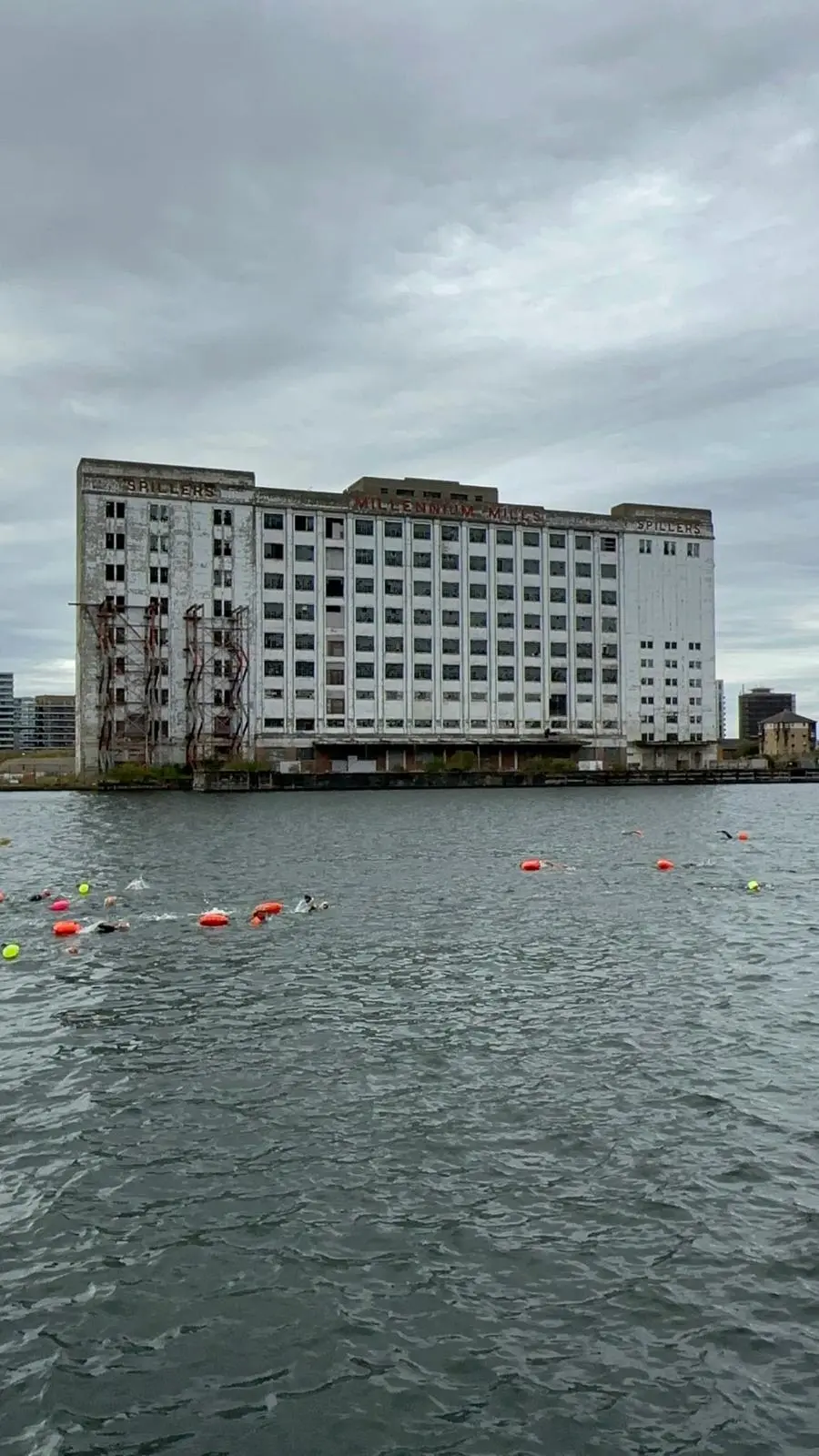 Grey dock water in front of the Millennium Mills, a string of orange and yellow tow floats marking swimmers in the distance.