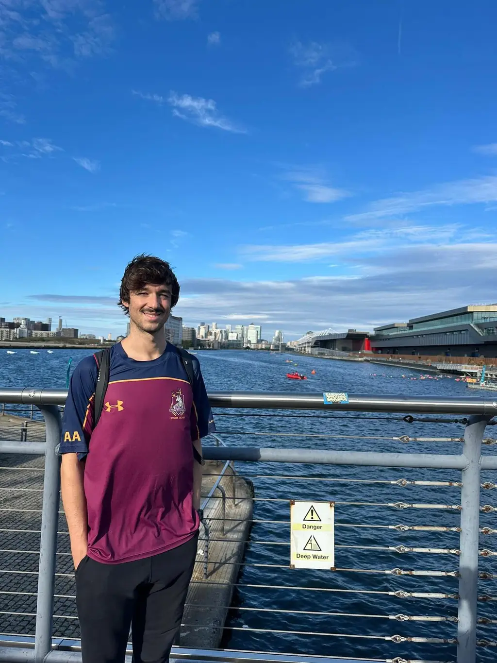 A person in a club polo shirt leaning on a dock railing under a clear sky, the water and the far bank behind, before the swim.