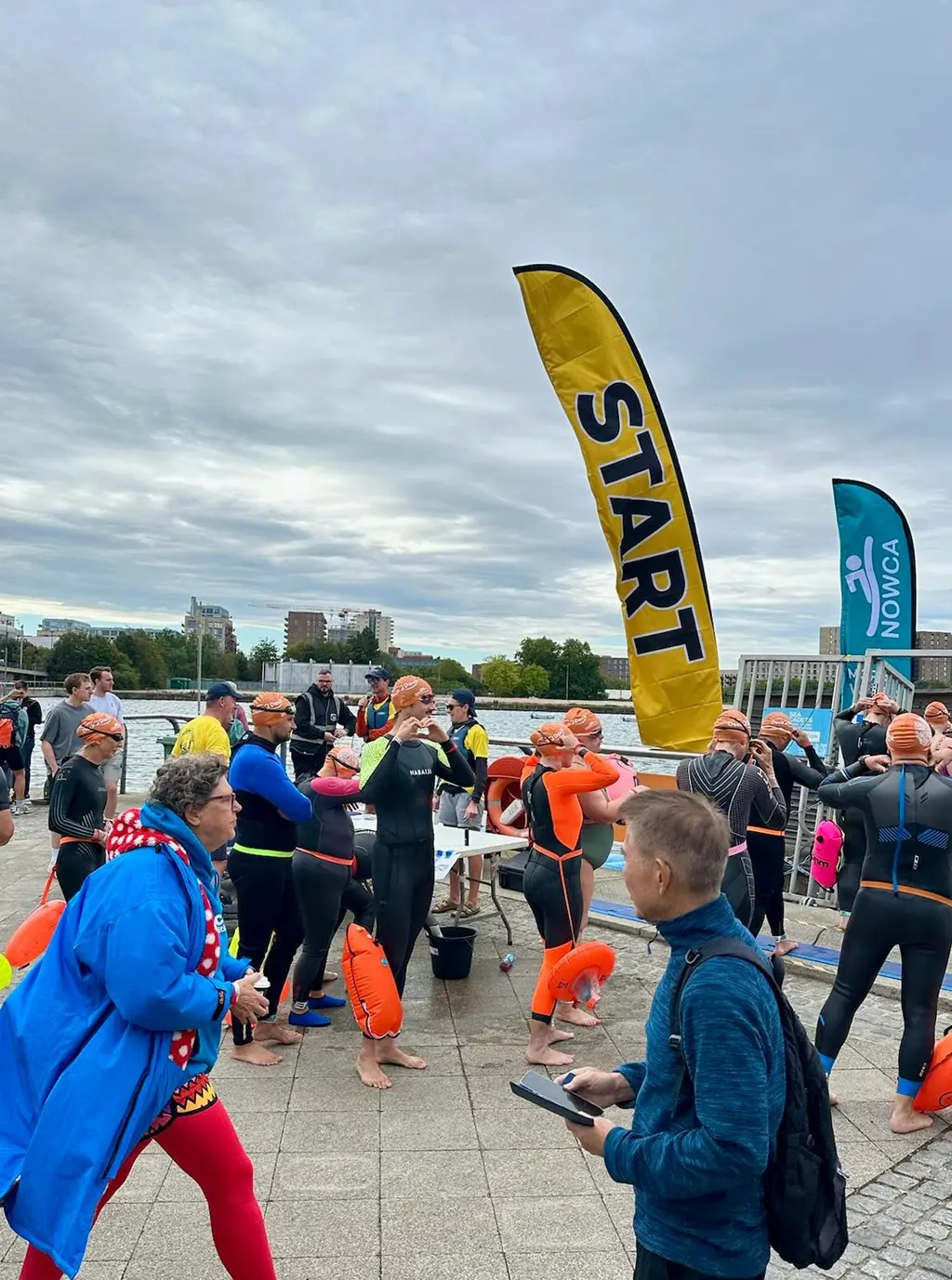 A yellow start flag on the dockside, swimmers in orange caps filing towards the water past a marshal.