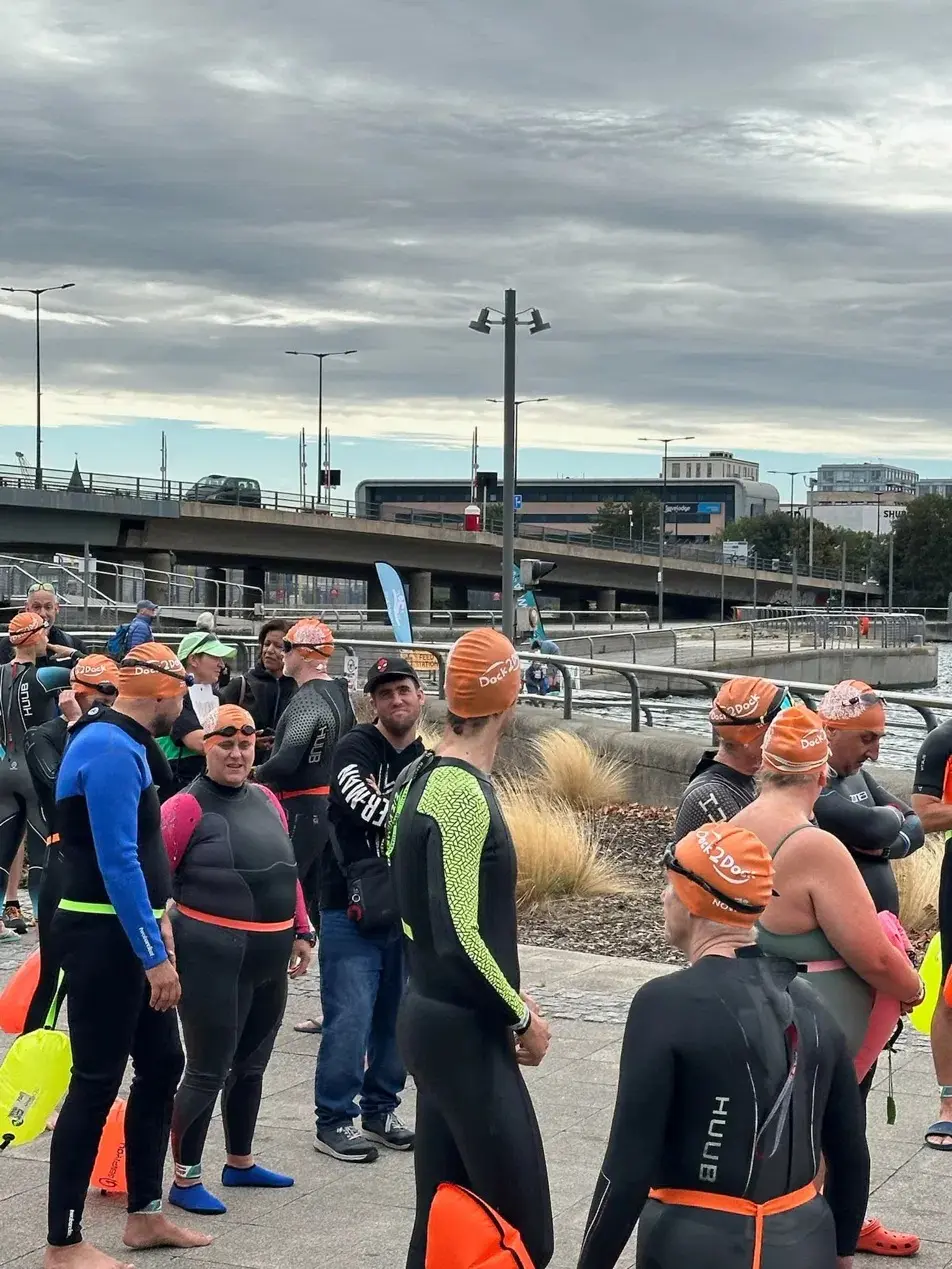 A crowd of swimmers in wetsuits and orange caps standing on the dockside waiting, a road bridge behind them.